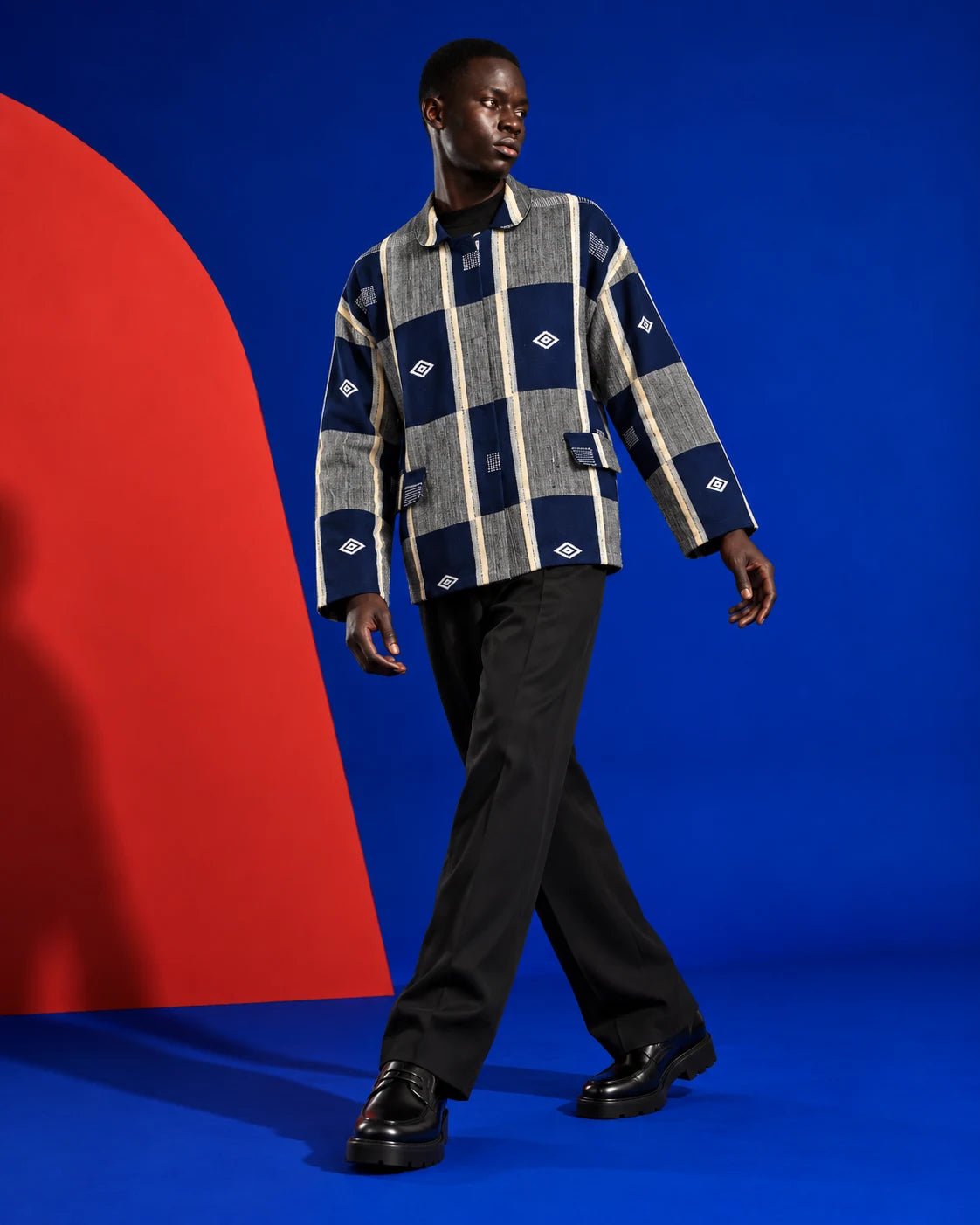 Veste boule Sénoufo bleu marine et gris portée fermée par un homme en pantalon noir, sur un fond bleu et rouge.