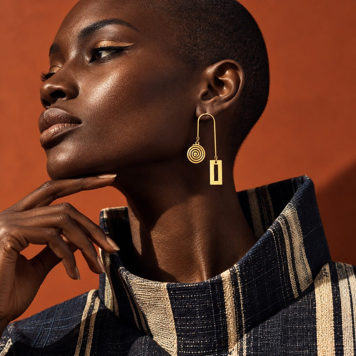Mannequin portant les boucles d'oreilles graphiques Anta de la collection Ligne, en acier inoxydable doré, avec un col en pagne tissé sur fond terracotta – Kaolack Créations