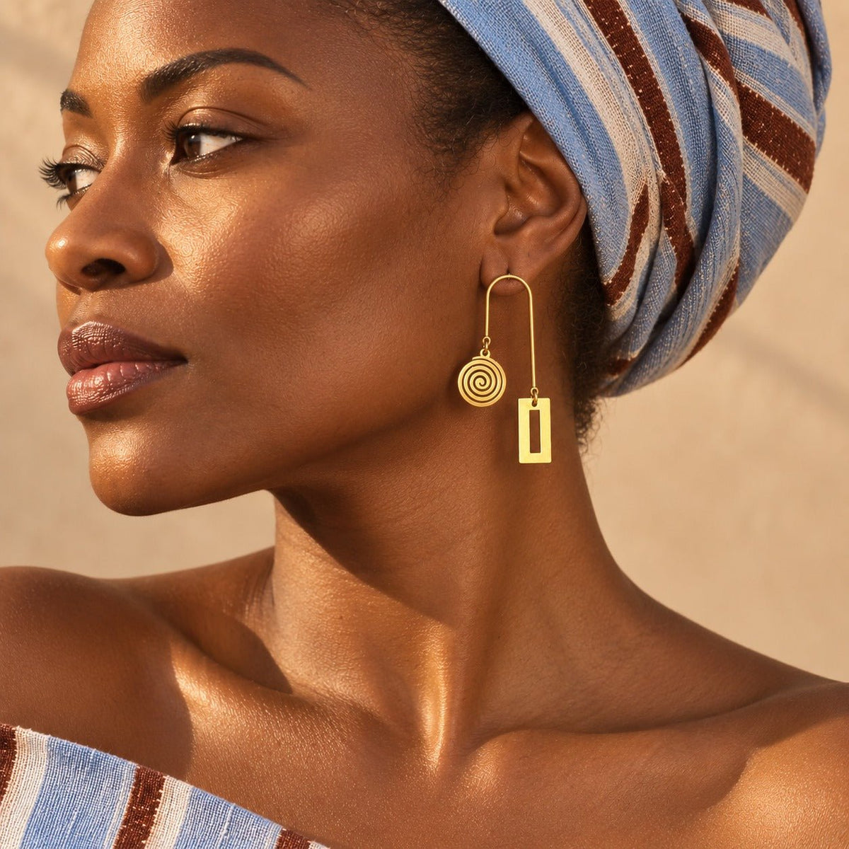 Femme portant les boucles d'oreilles graphiques Anta de la collection Ligne, en acier inoxydable doré, avec un foulard en pagne tissé – Kaolack Créations