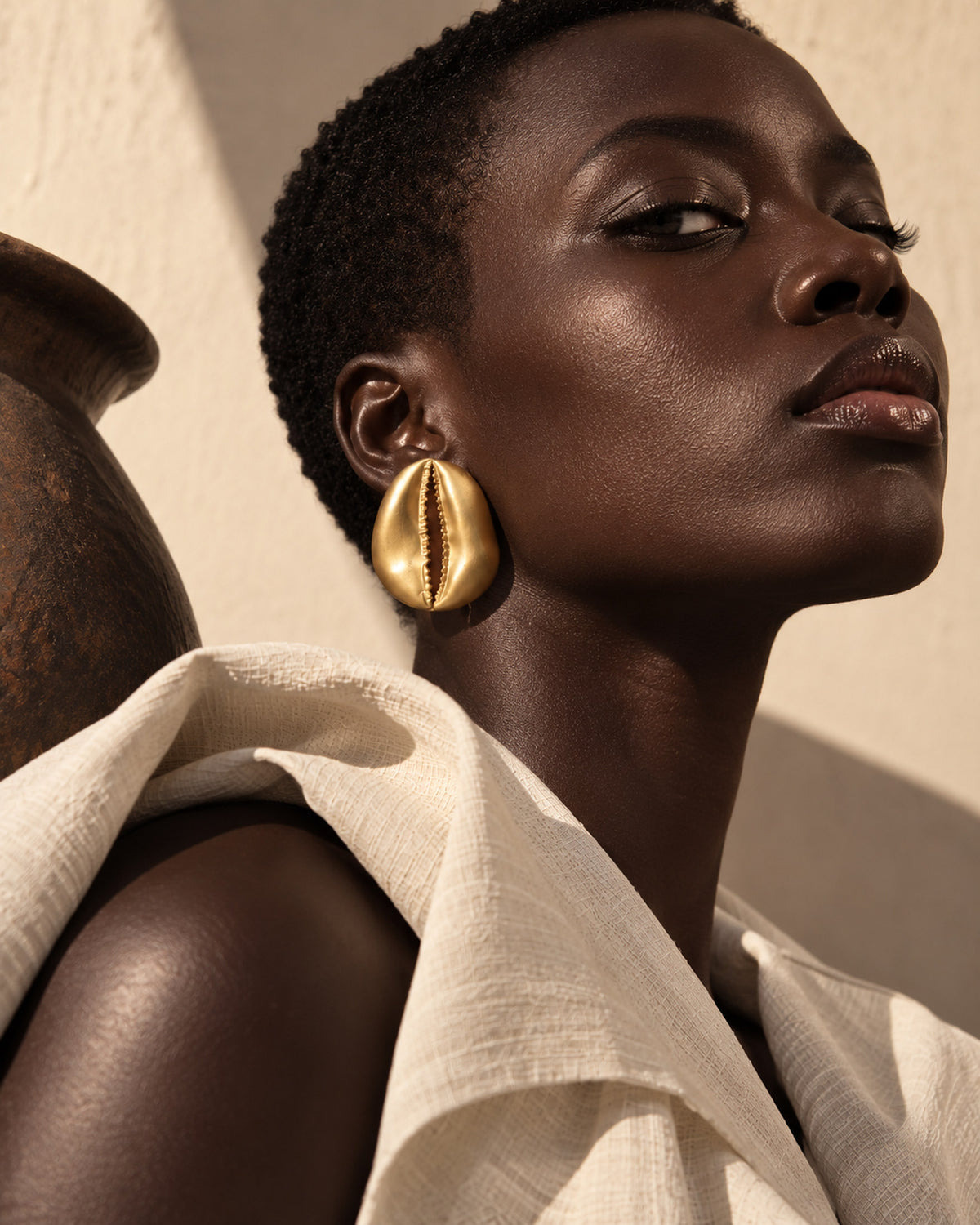 Grande boucle d'oreille cauri en bronze portée de profil, cheveux courts et drapé écru