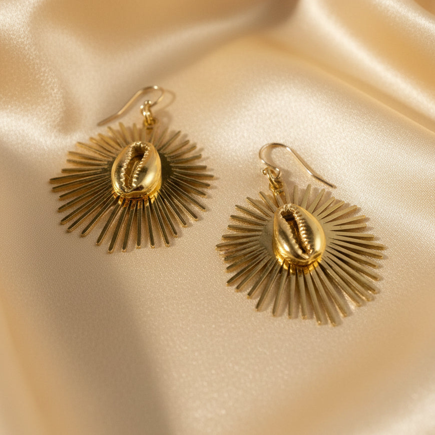 Boucles d’oreilles dorées avec cauri central et rayons solaires, bijou africain artisanal Kaolack Créations, photographié sur fond de soie glamour
