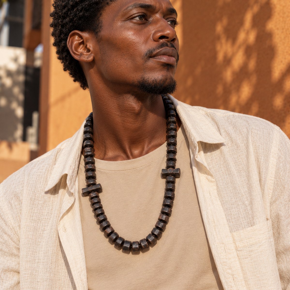 Collier long Guele Djiri en perles de bois porté sur une chemise en lin ouverte devant un mur ocre
