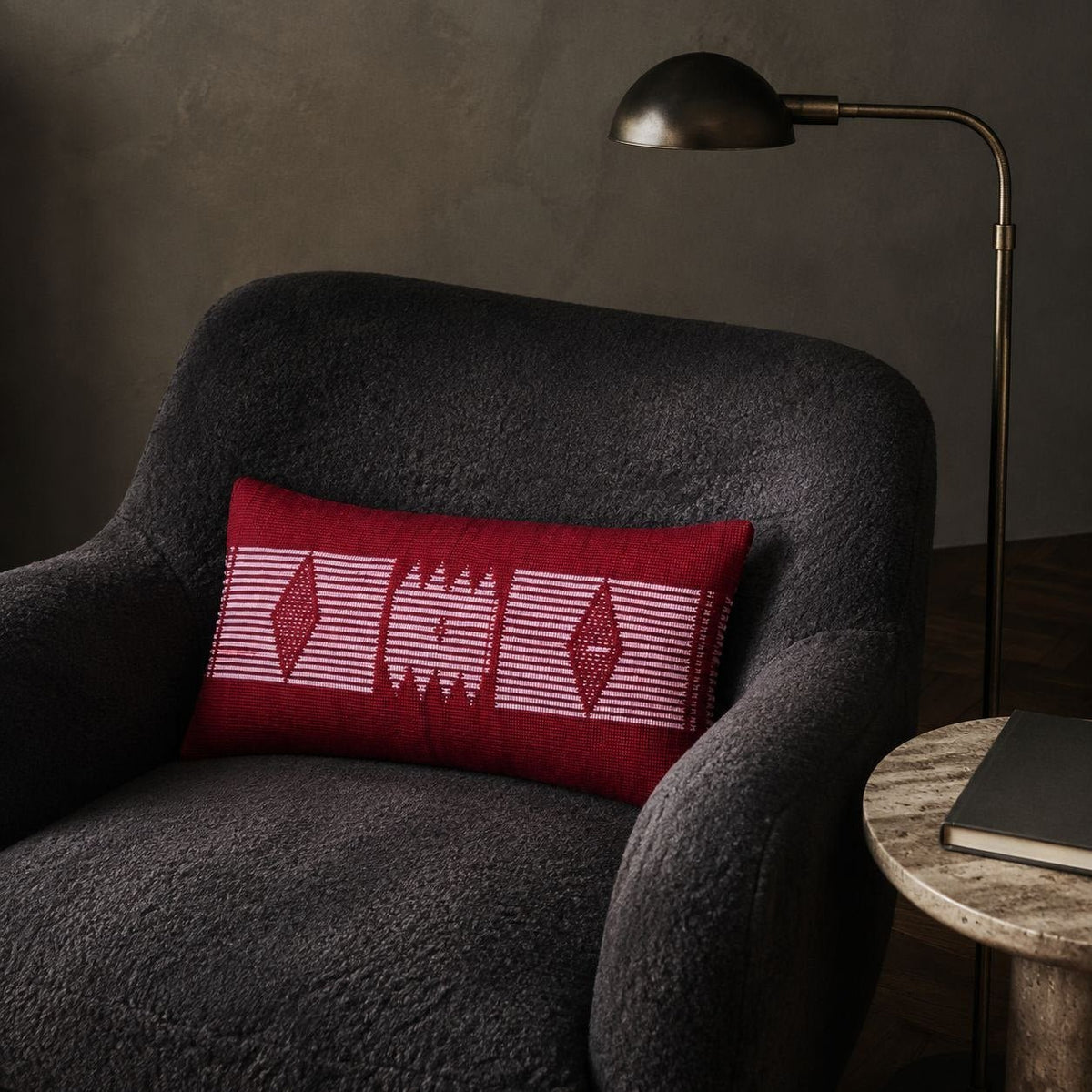 Housse de coussin Manjak rouge foncé sur un fauteuil en bouclé anthracite sous une lampe